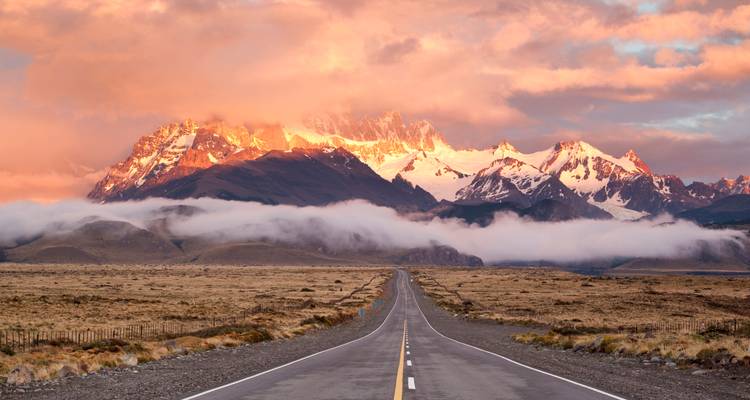Carretera recta y vacía que se dirige hacia picos nevados resplandecientes al amanecer con nubes bajas extendidas sobre el valle.