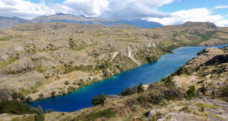 Diepblauwe rivier die zich slingerend door robuuste Patagonische heuvels onder verspreide wolken beweegt.