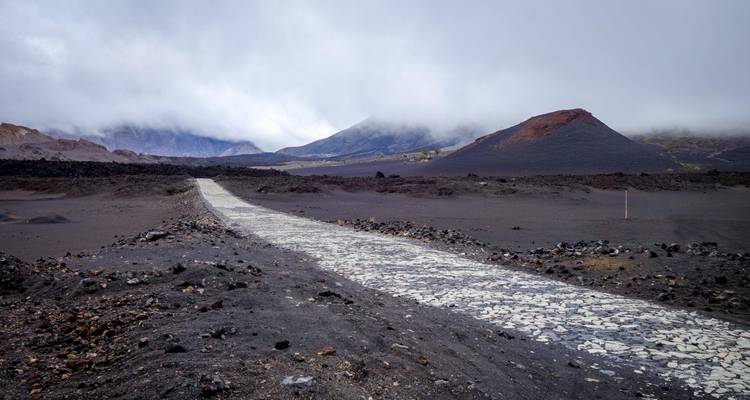 Desolate lava field with stone path leading toward mist-covered volcanic cones under grey skies.