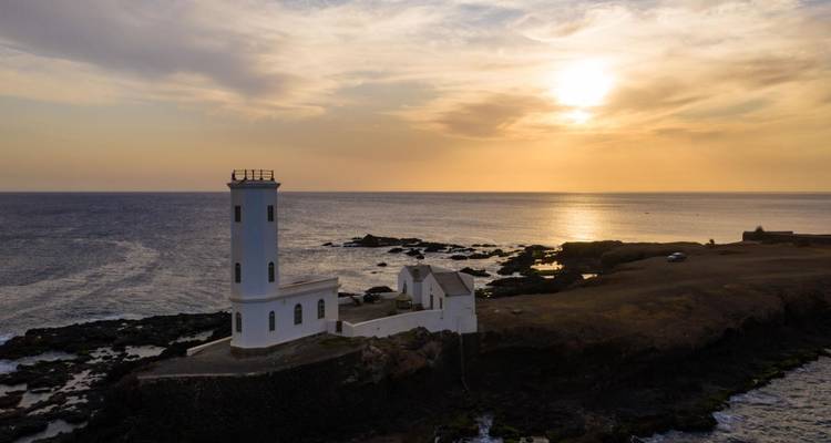 A white lighthouse and small keeper’s house stand on a rocky peninsula overlooking the sea beneath a golden sunset sky.