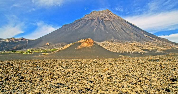 A dramatic dark-sloped volcano rises above a rough field of black lava rock under a bright blue sky.