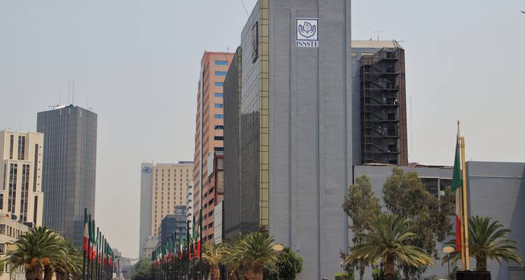 Modern office buildings with ISSSTE logo and avenue lined with flags and palm trees.