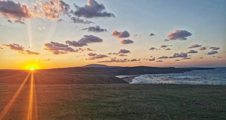 The sun sets over grassy headlands and a distant beach, colouring scattered clouds with warm hues.