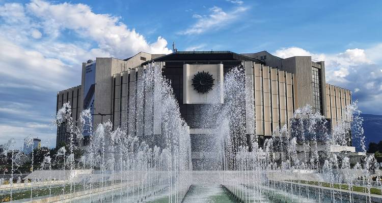 Modernist National Palace of Culture building fronted by high fountains spraying water under a blue sky.