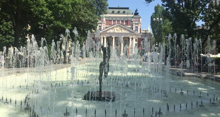 A long shallow fountain with multiple water jets leads the eye toward an ornate theatre framed by leafy trees.