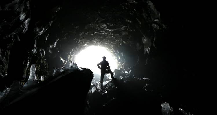 Silhouette of adventurer standing in icy lava tube cave entrance with bright backlight.