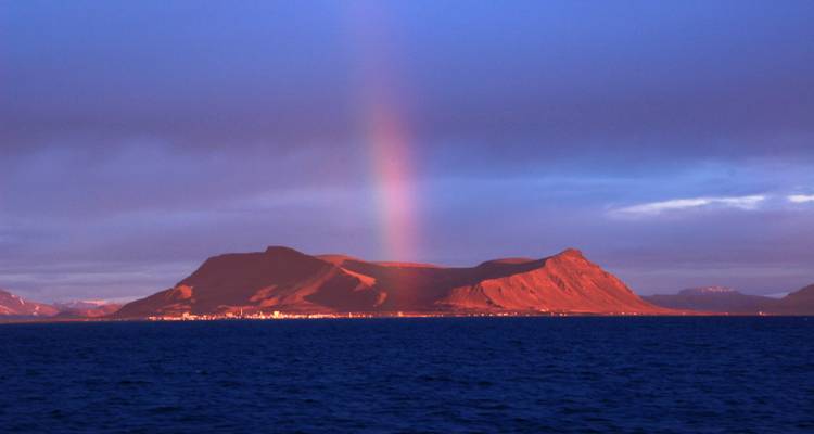 Isolated volcanic island glowing in warm light with a vivid rainbow rising into dramatic purple-blue evening clouds above a dark sea