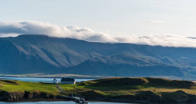 Serene coastal farmhouse on a green peninsula with dramatic cloud-capped mountains rising across the fjord in soft morning light