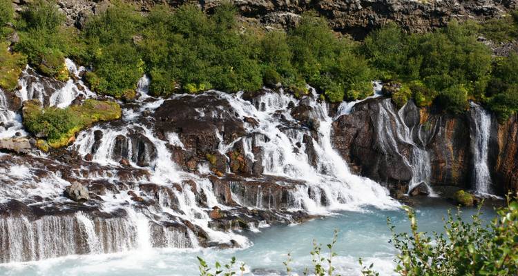 Multi-tiered waterfall cascading over dark volcanic rock into turquoise water surrounded by lush green foliage