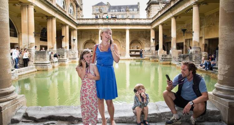 Famille souriante utilisant des audioguides à côté du bassin fumant des thermes romains à l'intérieur d'un complexe en pierre ancienne