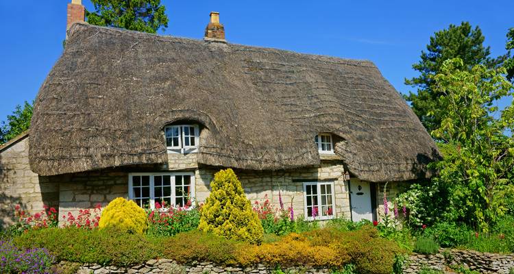 Charmante chaumière en pierre au toit de chaume entourée d'un jardin de cottage coloré sous un ciel bleu éclatant dans les Cotswolds