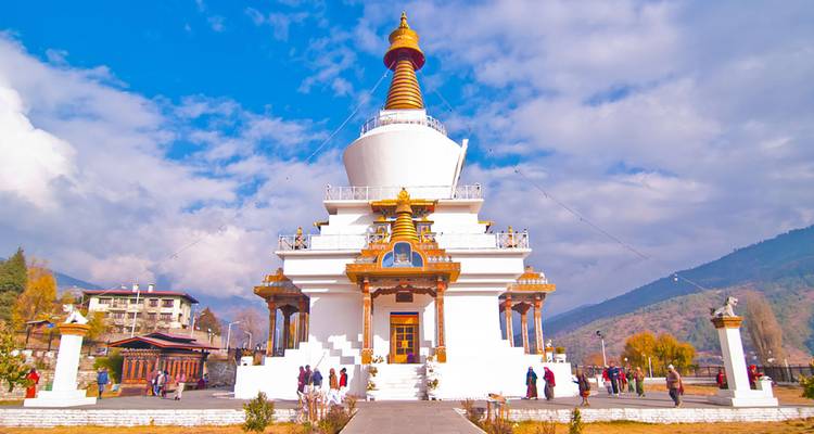 Chorten commémoratif blanc avec flèche dorée brille sous un ciel bleu éclatant, visiteurs à la base.