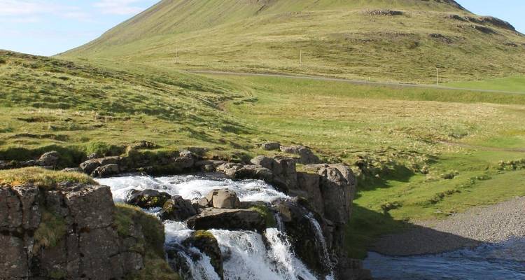 Small waterfall tumbling over rocks with grassy Kirkjufell mountain rising behind.