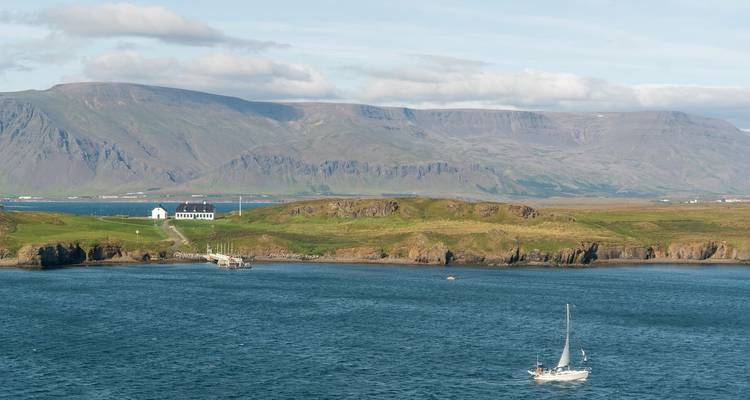 Sailboat on blue waters with rugged Icelandic coastline and mountains in the distance.