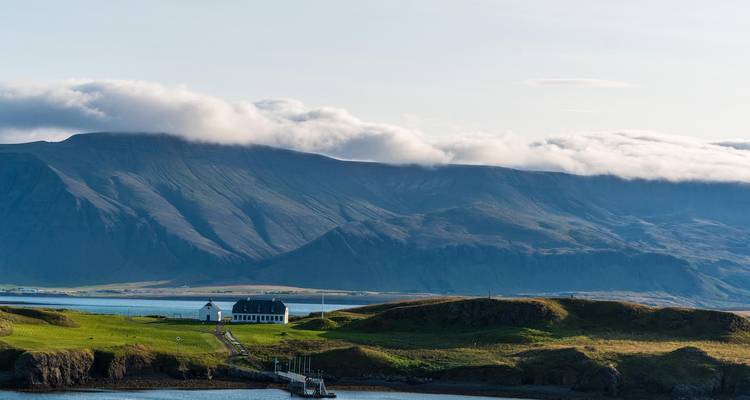 White cottage beside a pier with dramatic cloud-capped mountains beyond.