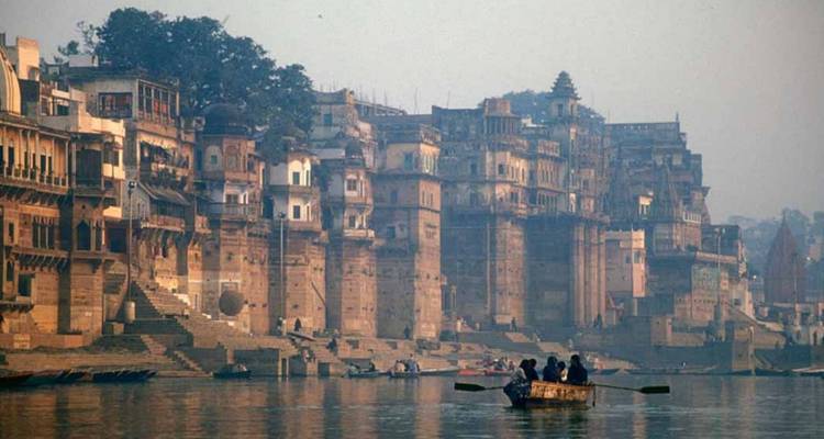 Dunstige Morgenansicht der Ghats von Varanasi mit historischen Ufergebäuden, die sich über dem Ganges erheben, und kleinen Booten auf dem Wasser.