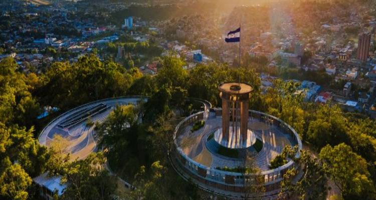 Monument au sommet d'une colline avec un grand drapeau hondurien surplombant une ville tentaculaire baignée dans la lumière dorée du matin.