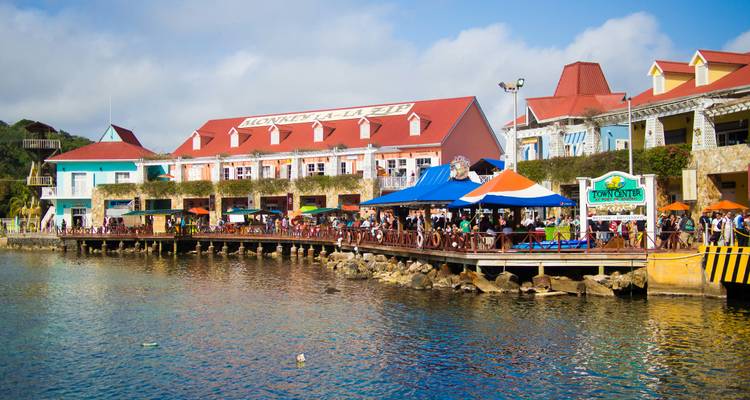 Village commerçant coloré au bord de l'eau avec une ambiance de port de croisière se reflétant dans l'eau bleue calme sous un ciel lumineux.