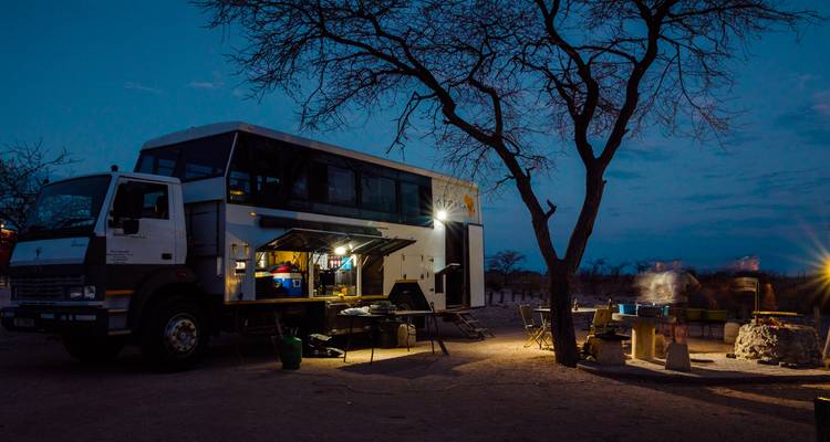 Camion d'expédition terrestre éclairé par des lanternes dans un campement isolé sous un ciel crépusculaire près d'un arbre dénudé.