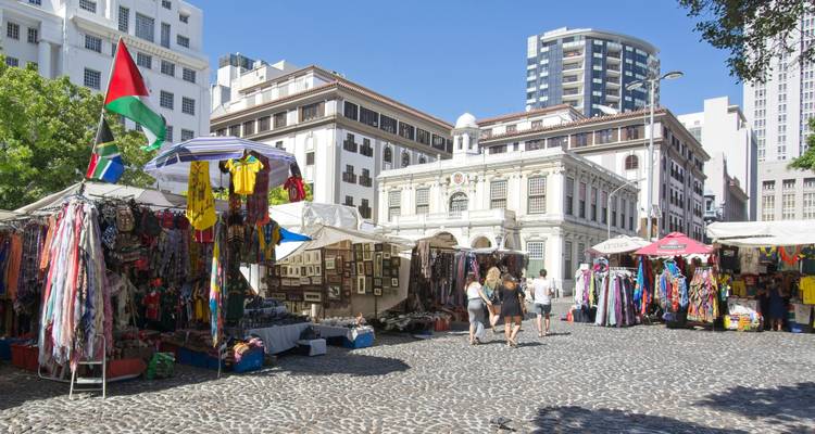 Mercado callejero al aire libre con puestos de telas coloridas y compradores en una plaza empedrada rodeada de edificios coloniales.
