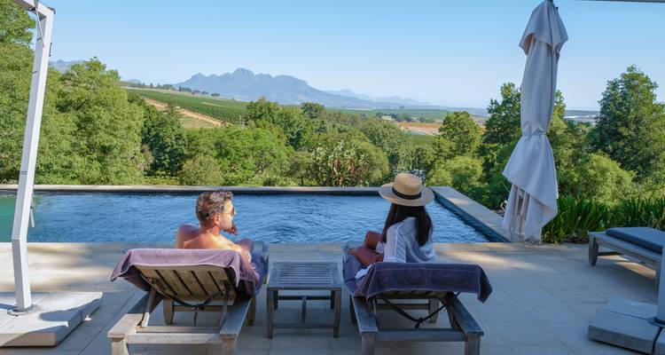 Pareja relajándose en tumbonas junto a la piscina con vista a viñedos exuberantes y una cordillera distante.