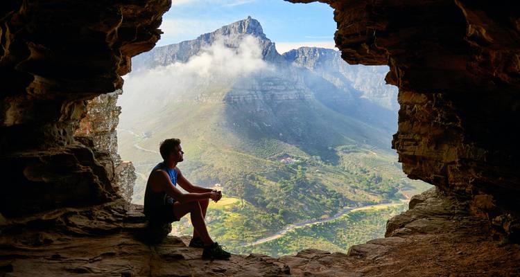 Excursionista sentado dentro de una abertura de cueva rocosa, admirando un paisaje montañoso dramático parcialmente cubierto por nubes.