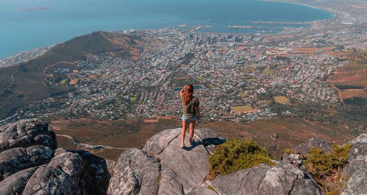 Una mujer se encuentra en una repisa rocosa en la cima de una montaña mirando Ciudad del Cabo y la costa muy abajo.