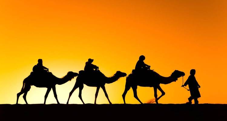 Silhouettes de cavaliers sur des chameaux menés par un guide traversant une dune de sable sous un coucher de soleil orange éclatant.