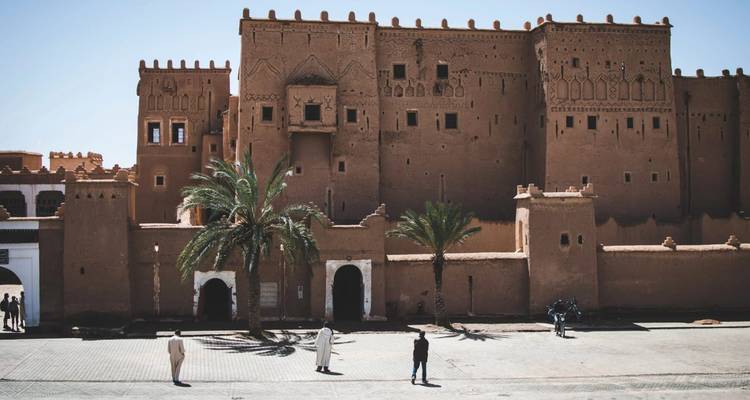 Des visiteurs se tiennent devant une grande kasbah couleur sable avec des palmiers bordant la cour ensoleillée.