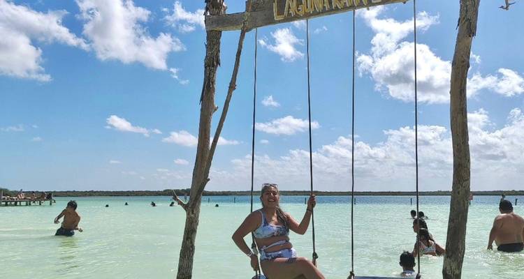 Jeune femme souriante sur une balançoire en bois installée au-dessus de l'eau turquoise d'un lagon pendant que des nageurs profitent des eaux peu profondes.