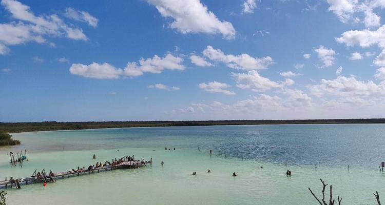 Scène de large lagon avec des eaux peu profondes turquoise pâle, une jetée en bois remplie de visiteurs, et des nageurs dispersés dans l'eau.