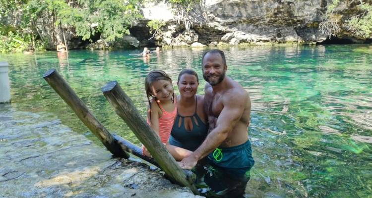 Famille souriante posant sur des marches en bois dans un cénote d'eau douce entouré d'un feuillage luxuriant et d'une eau émeraude.