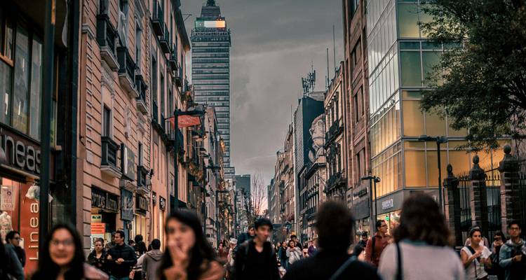 Überfüllte Fußgängerstraße in der Innenstadt von Mexiko-Stadt, gesäumt von historischen Gebäuden und modernen Geschäften in der Abenddämmerung.