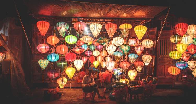 Étal de marché de nuit illuminé de dizaines de lanternes colorées tandis que deux vendeurs se détendent en dessous.