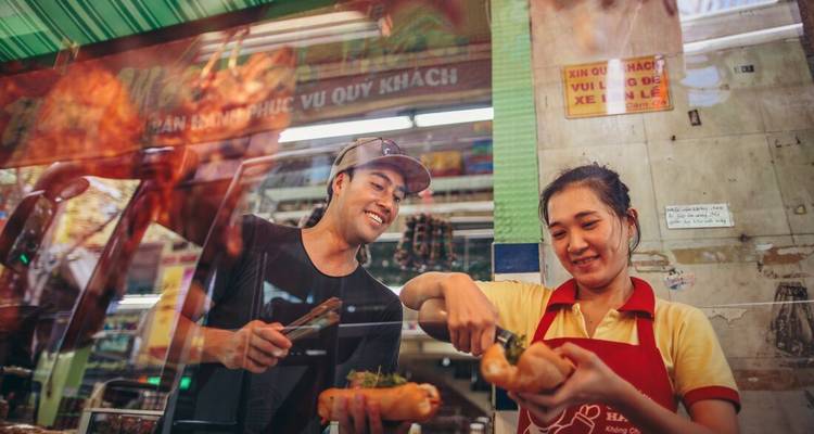 Les vendeurs de nourriture de rue rient en préparant des banh mi frais derrière un comptoir en verre.