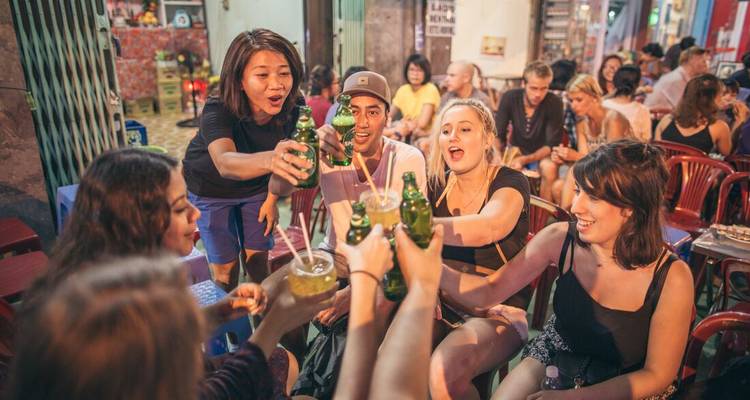 Un groupe d'amis lèvent leurs bouteilles dans un bar animé en terrasse la nuit.