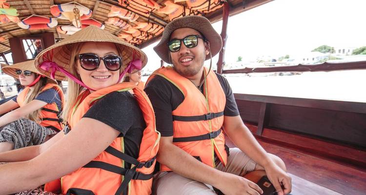 Des voyageurs coiffés de chapeaux coniques et vêtus de gilets de sauvetage orange sont assis à bord d'un bateau en bois à ciel ouvert.