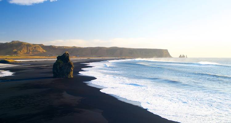 Golven rollen aan op een uitgestrekt zwart zandstrand met dramatische zeerotsen voor de zuidkust van IJsland.