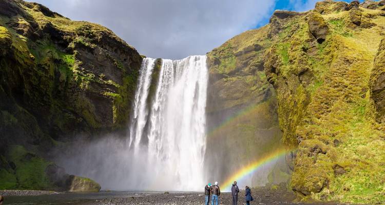 Skogafoss dondert in een bemoste kloof, dubbele regenbogen buigen terwijl toeschouwers aan de grindbasis staan.