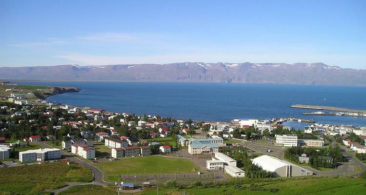 Ville côtière islandaise avec port et montagnes enneigées au loin sous un ciel bleu dégagé.
