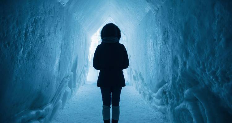 Visiteur en silhouette dans un tunnel de glace éclairé en bleu marchant vers une sortie lumineuse.