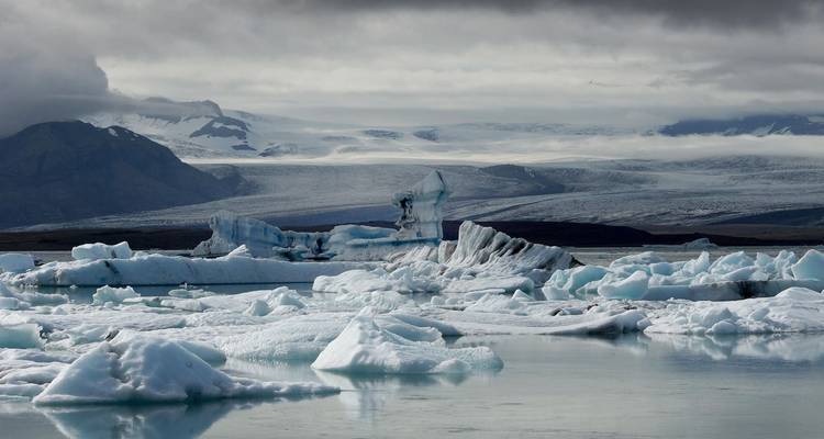 Moody skies hang over an icy lagoon dotted with angular blue icebergs and distant glacier tongues.