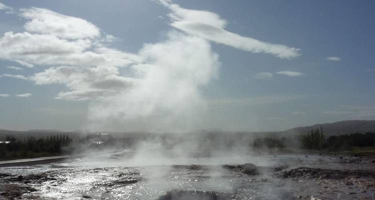 A plume of boiling water and steam erupts from a geyser field under bright skies.