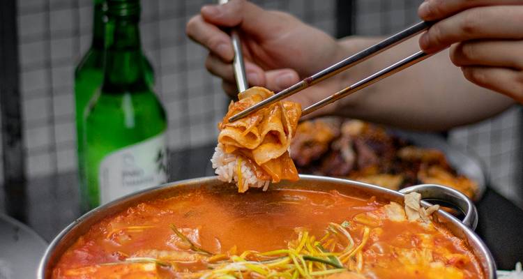 Los palillos en primer plano levantan arroz y kimchi de una olla de estofado coreano burbujeante junto a una botella verde de soju.