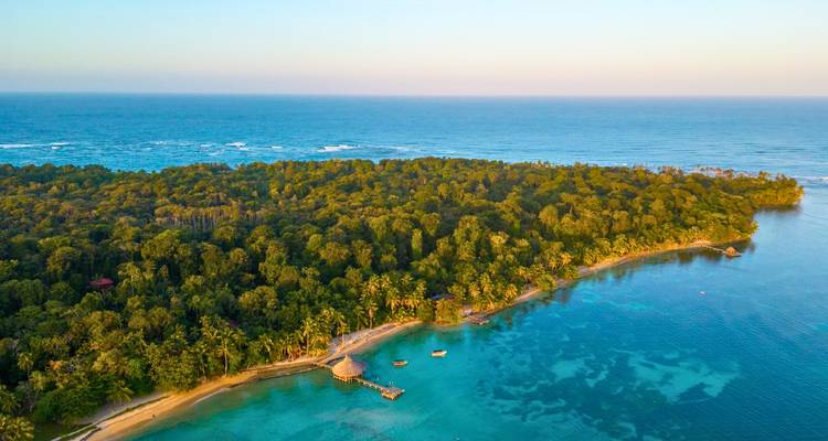 Dronevizier van een weelderig tropisch eiland omzoomd door turquoise water en koraalriffen, met open zee daarachter onder zacht licht van de late dag.