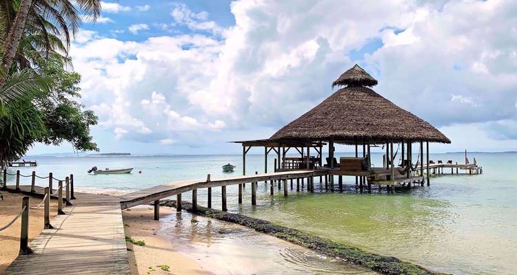 Idyllisch rieten paviljoen op een houten steiger die zich uitstrekt over kalm turquoise water met palmbomen en een met wolken bezaaide hemel.