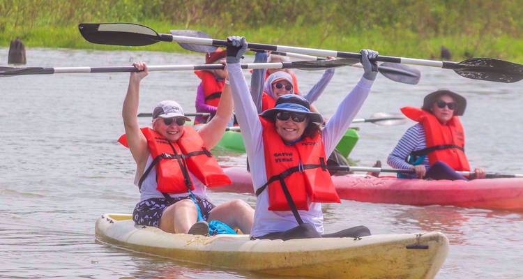 Enthousiaste kajakkers met rode zwemvesten peddelen samen op kalm water, een van hen steekt de peddels triomfantelijk omhoog.