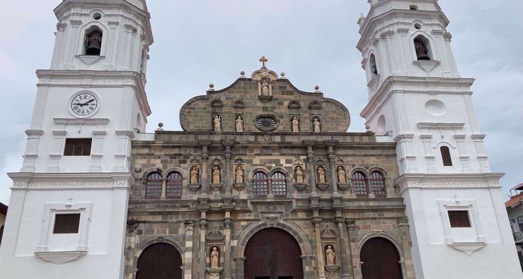 Voorgevel van een kathedraal met twee torens van steen en witgekalkte muren met klok en standbeelden onder een bewolkte hemel.