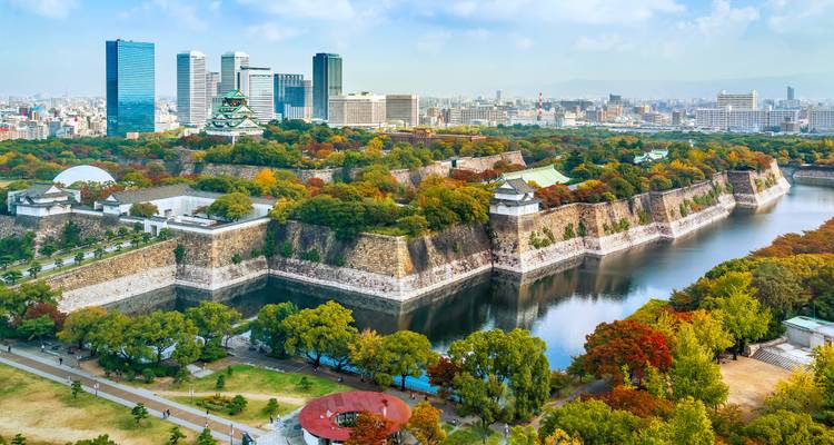 Luftpanorama der Steinwälle der Burg Osaka und des umliegenden Wassergrabens, eingerahmt von Herbstbäumen mit der modernen Skyline im Hintergrund.