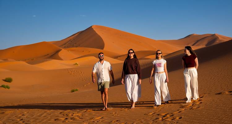 Quatre voyageurs marchent en file à travers d'imposantes dunes orangées sous un ciel désertique bleu et dégagé.